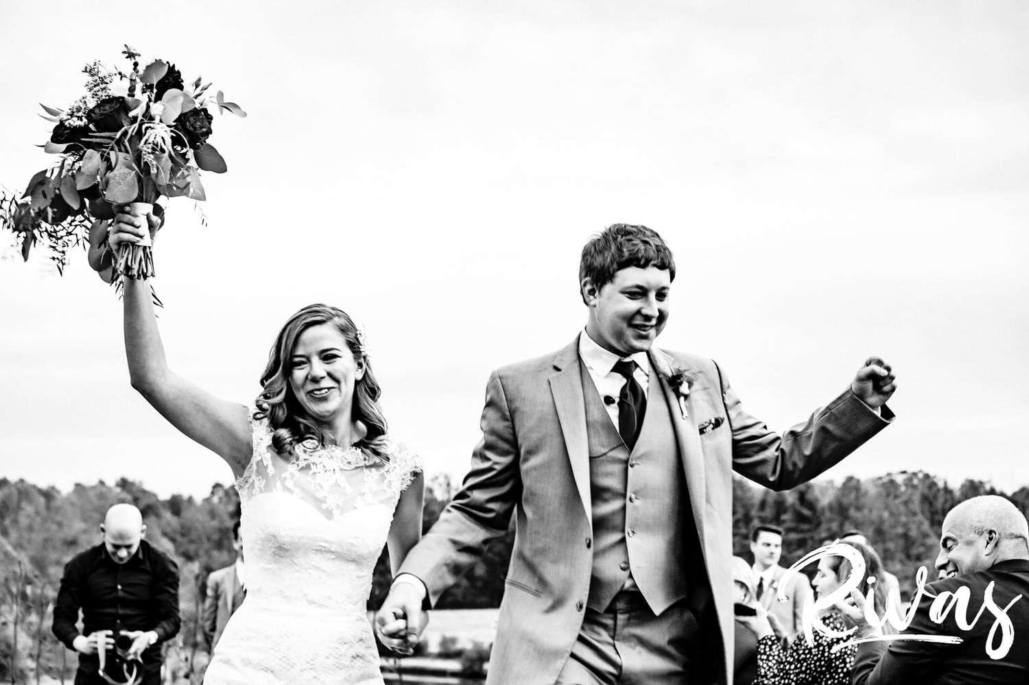 A candid black and white picture of a bride and groom throwing their arms up in celebration after their fall wedding ceremony. 