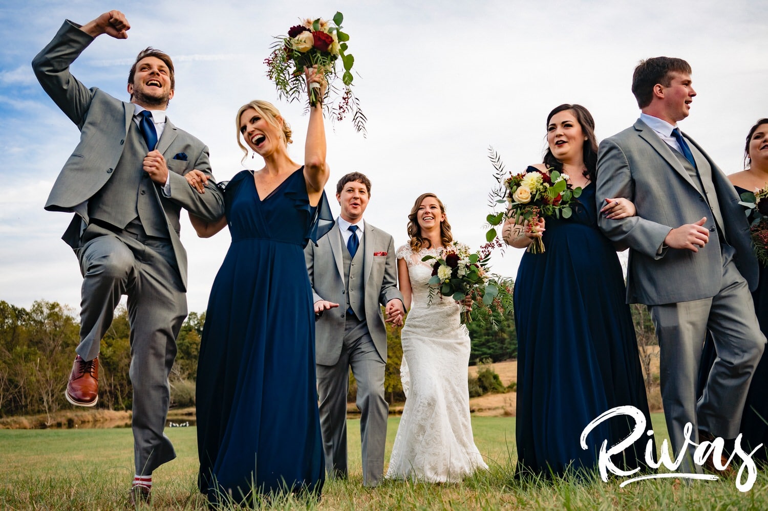 A causal, colorful picture of a bride and groom walking arm in arm with their joyous wedding party across a field on their fall wedding day at The Barns at Hamilton Station Vineyard. 