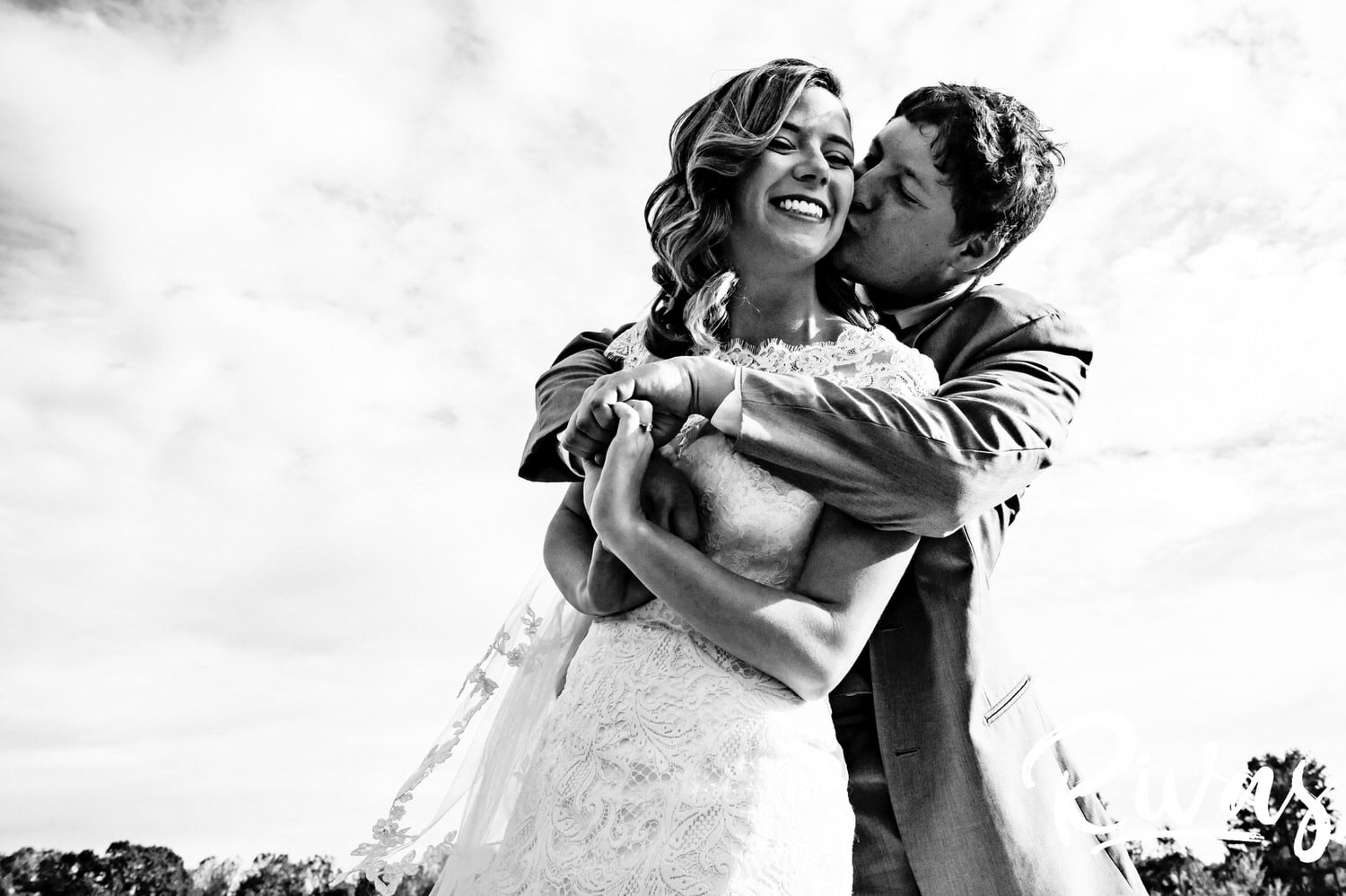 An up-close, black and white picture of a groom hugging his bride from behind as he kisses her cheek on the afternoon of their fall wedding day. 