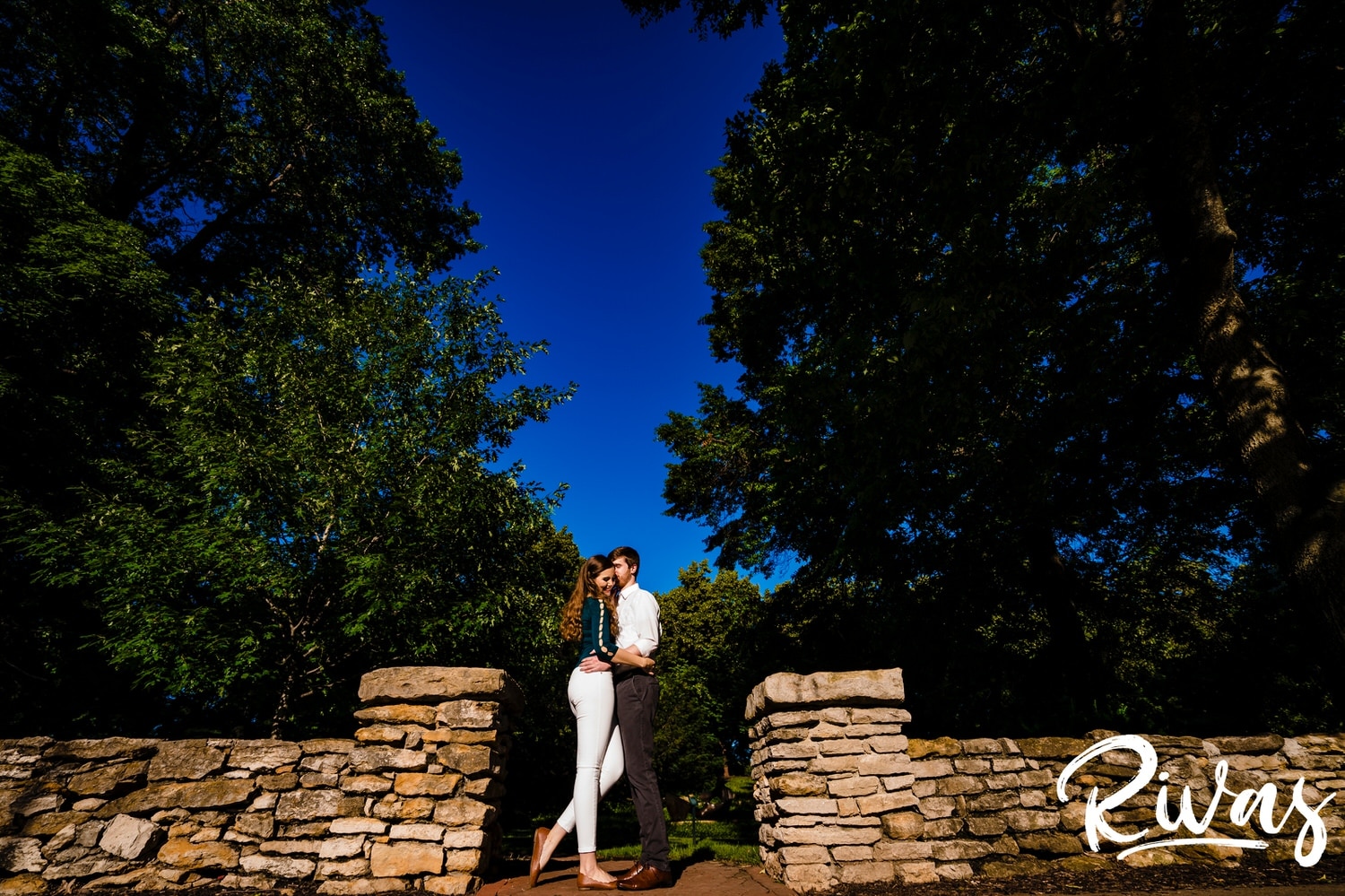 A vibrant portrait of an engaged couple sharing a kiss underneath a bright blue sky during their summer engagement session at The Nelson Atkins Museum of Art in Kansas City. 