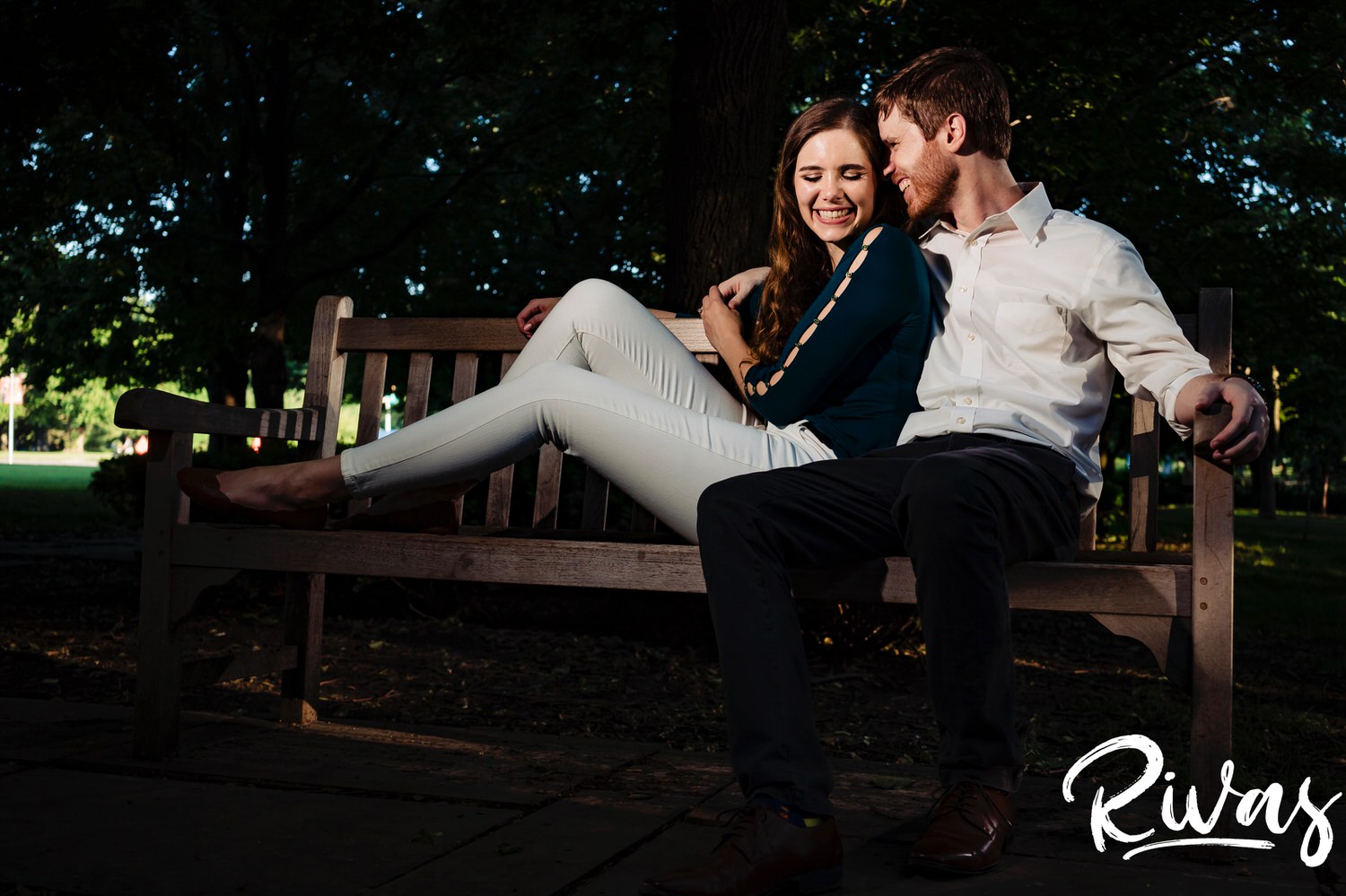 A relaxed portrait of an engaged couple sharing an embrace as they sit on a wooden bench surrounded by trees at The Nelson Atkins Museum of Art during their Kansas City Engagement Session.