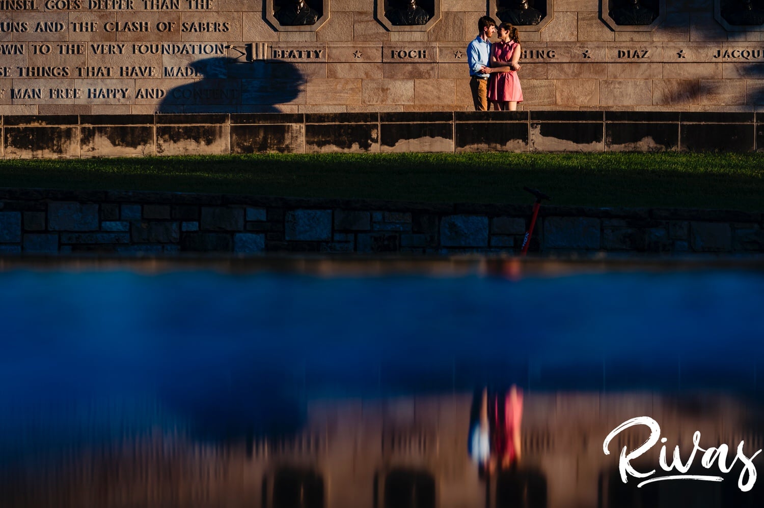 A wide portrait of a couple standing on a stone wall dancing, with their reflection visible in the fountain outside Union Station during their engagement session in Kansas City. 