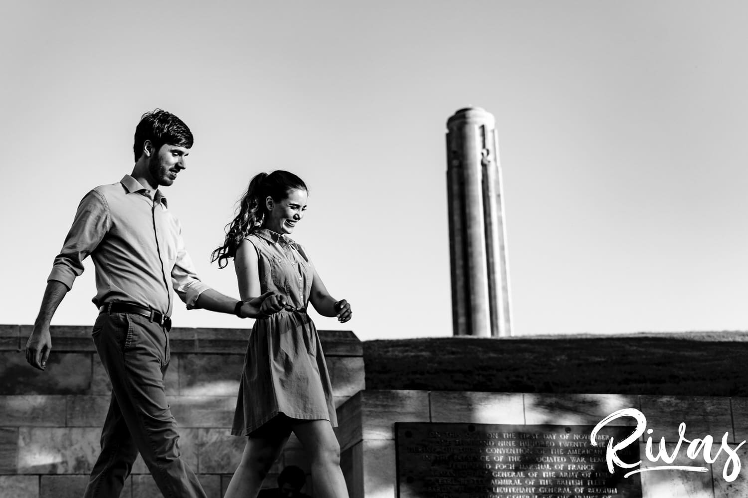 A candid black and white picture of an engaged couple walking hand-in-hand at the base of Liberty memorial during their Kansas City engagement session. 