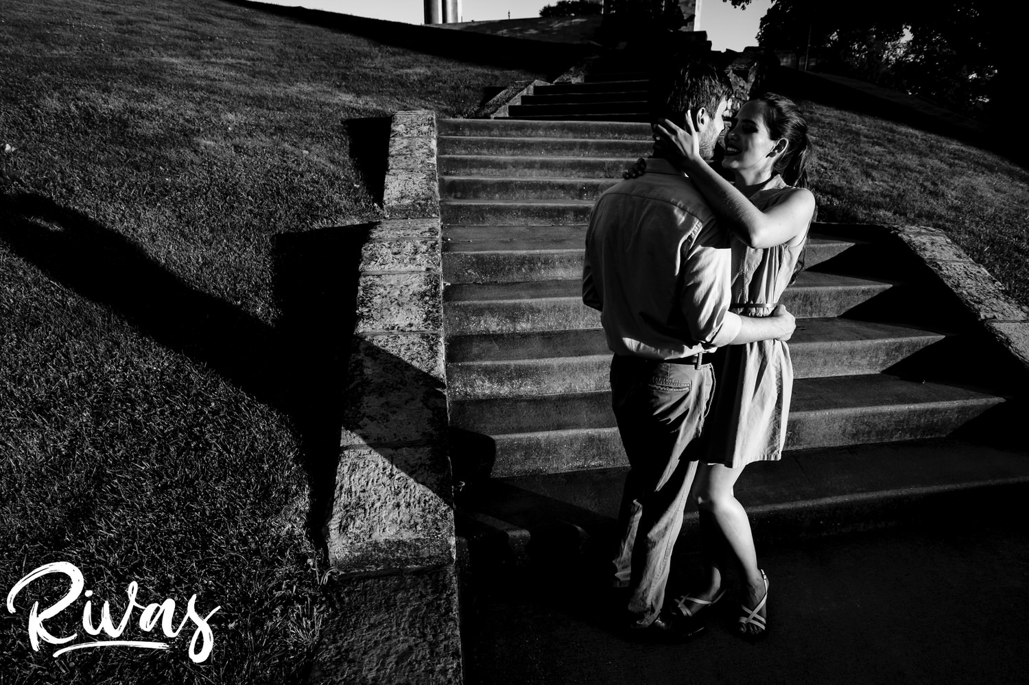 A playful black and white picture of an engaged couple stopping to share an embrace as they walk up a flight of stairs at the base of Liberty Memorial Park in Kansas City during their summer engagement session. 