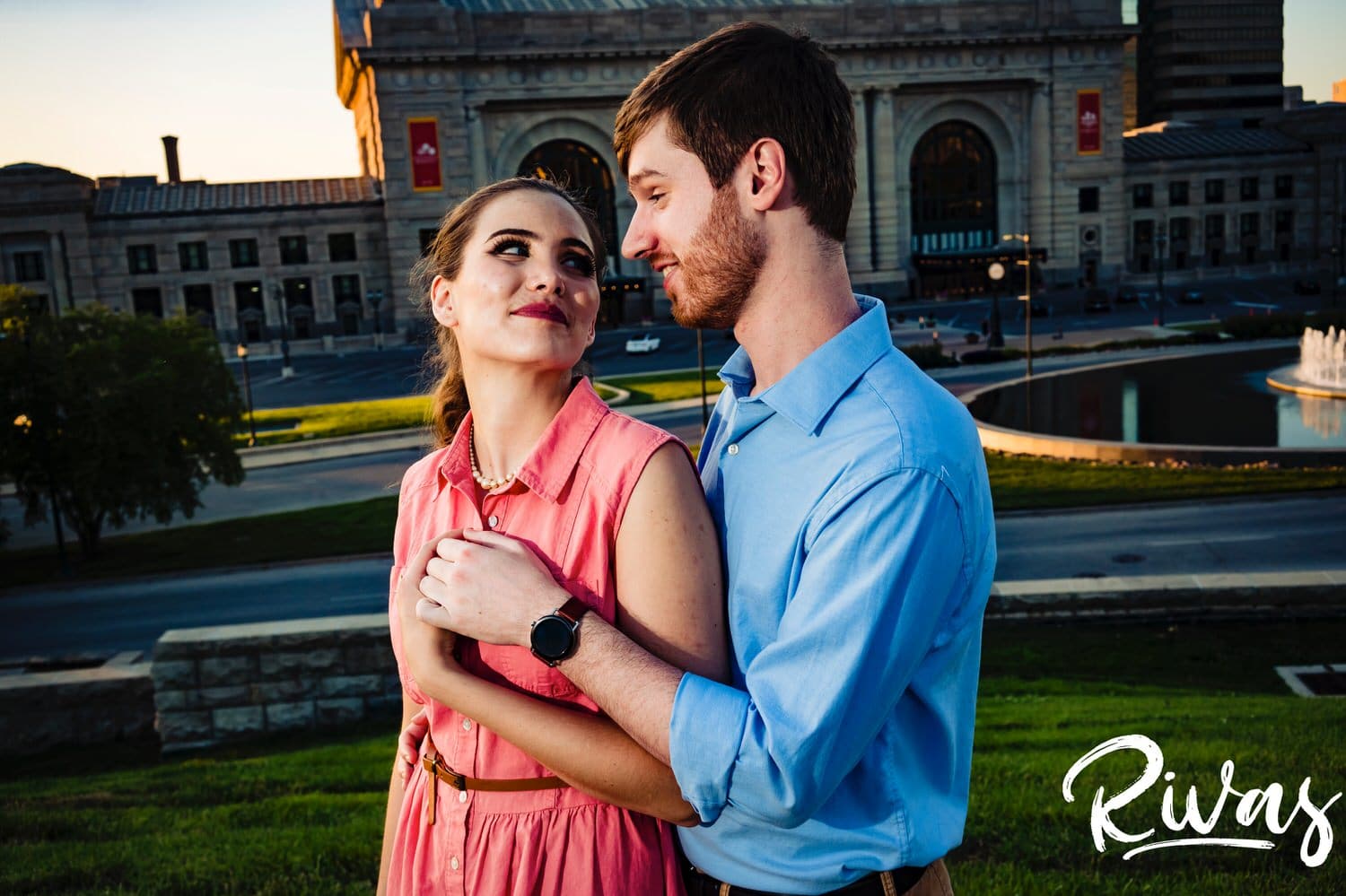A colorful, sweet picture of an engaged couple sharing an embrace as they stand on a stone wall in front of Union Station in Kansas City during their summer engagement session. 