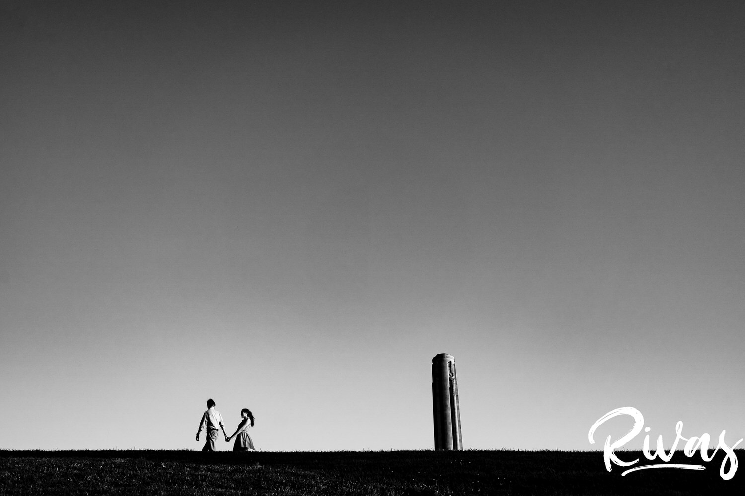 A very wide, black and white picture of a couple walking hand in hand in front of the base of Liberty Memorial in Kansas City during their summer engagement session. 