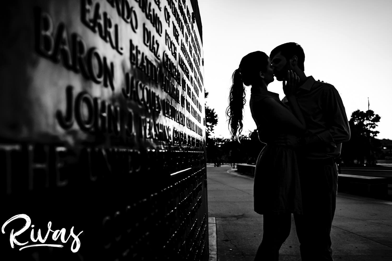 An intimate black and white portrait of an engaged couple sharing a kiss, silhouetted against the setting sun during their summer engagement session in Kansas City. 