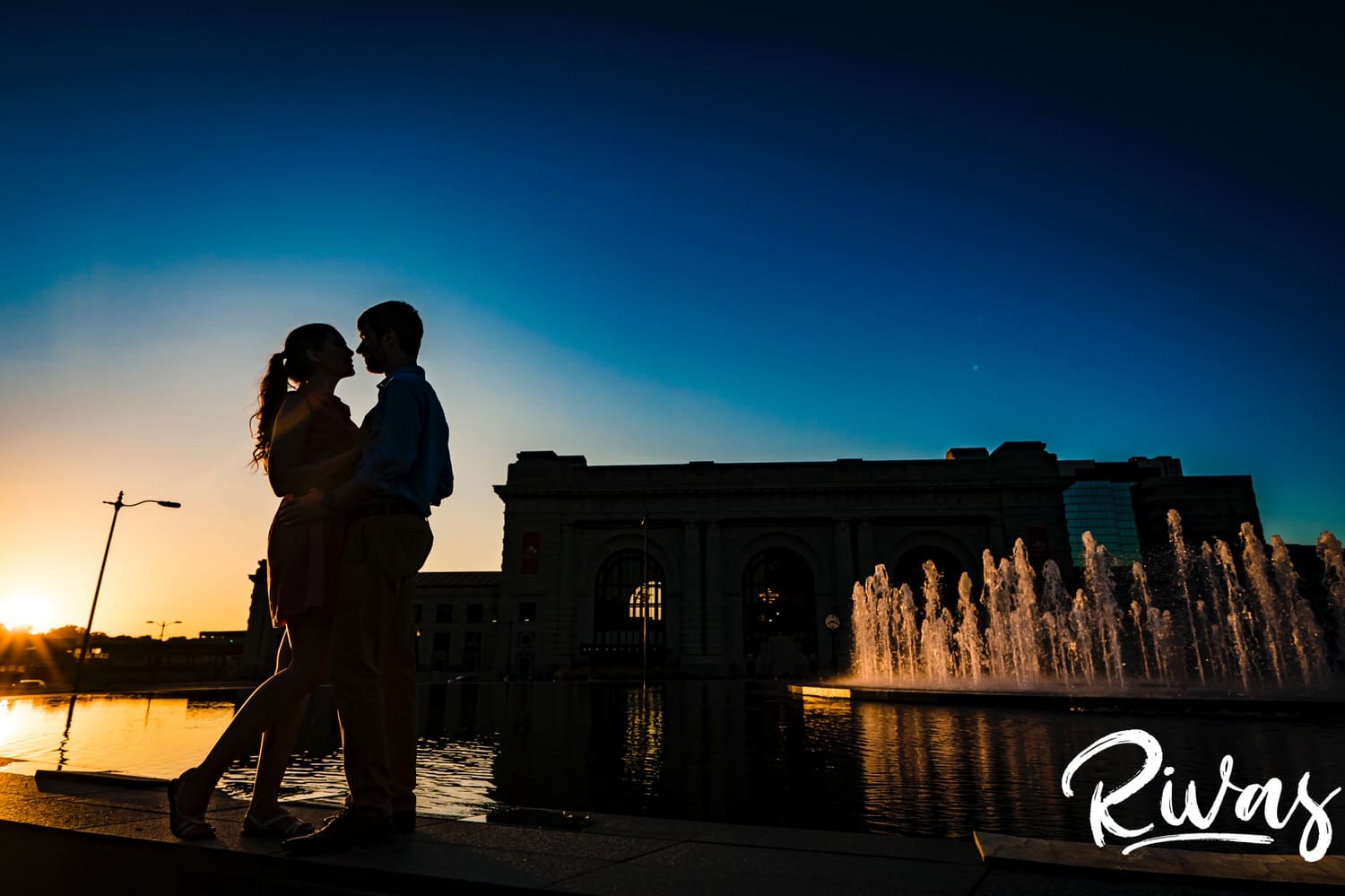 A vibrant silhouetted picture of an engaged couple sharing an embrace as they lean up against the edge of the fountain outside Kansas City's Union Station during their summer engagement session. 