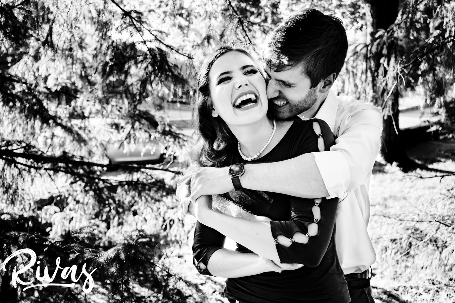 A candid black and white picture of a man hugging his fiance as she laughs hysterically during their summer engagement session at The Nelson Atkins Museum of Art in Kansas City.
