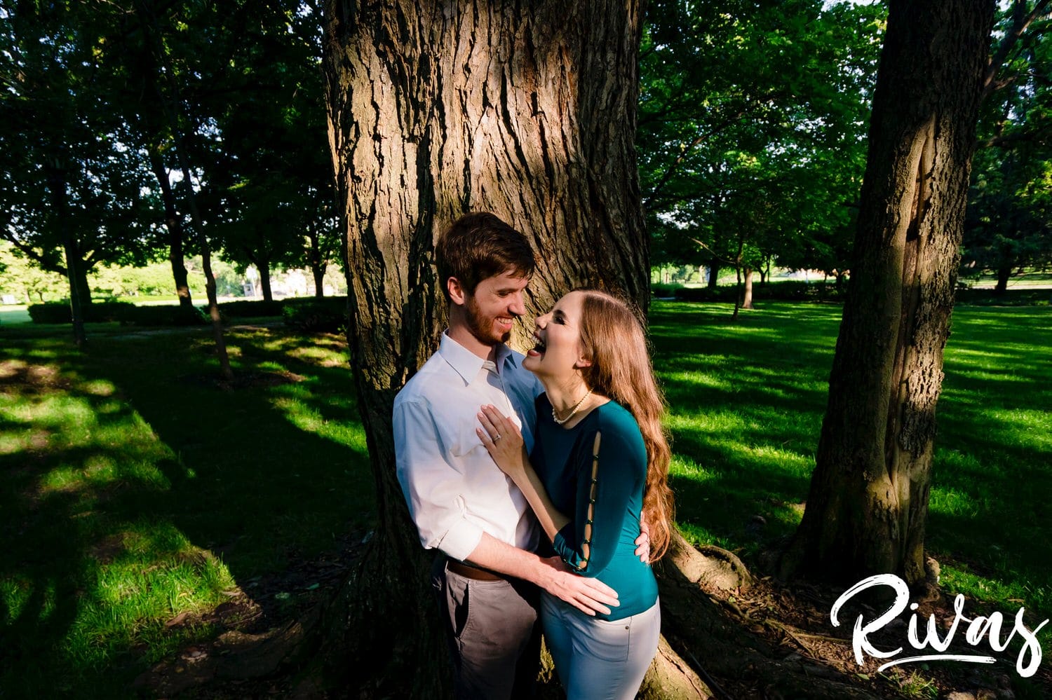 A close-up, candid picture of an engaged couple sharing an embrace and laughing together as they stand underneath a tree during their engagement session at The Nelson in Kansas City. 