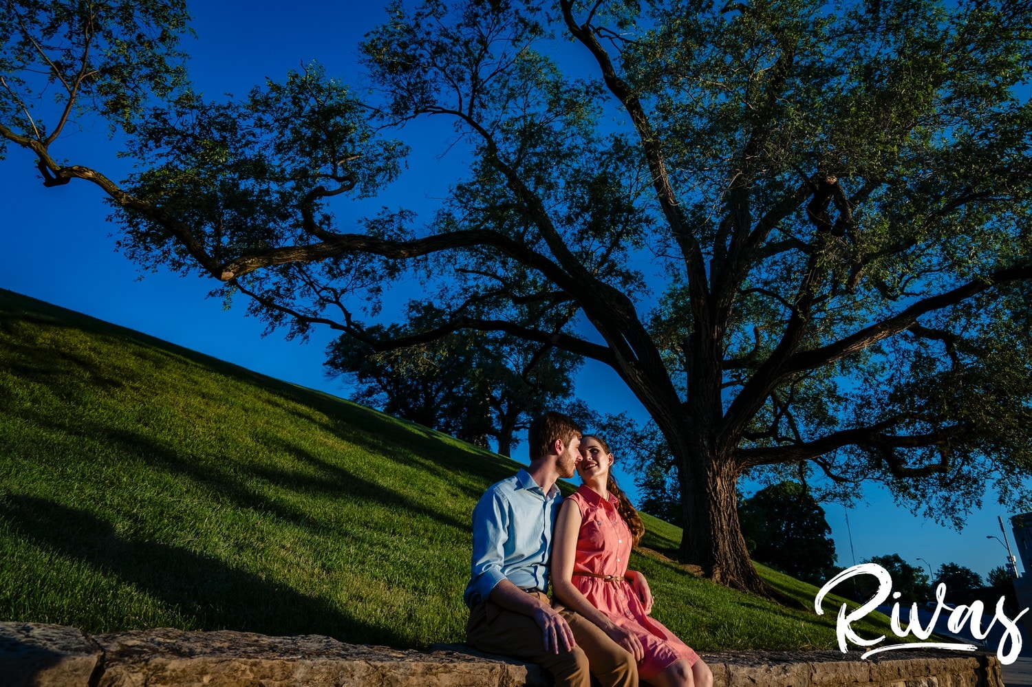 A colorful picture of an engaged couple sitting on a stone wall underneath a tree at the base of Liberty memorial Park during their KC summer engagement session. 