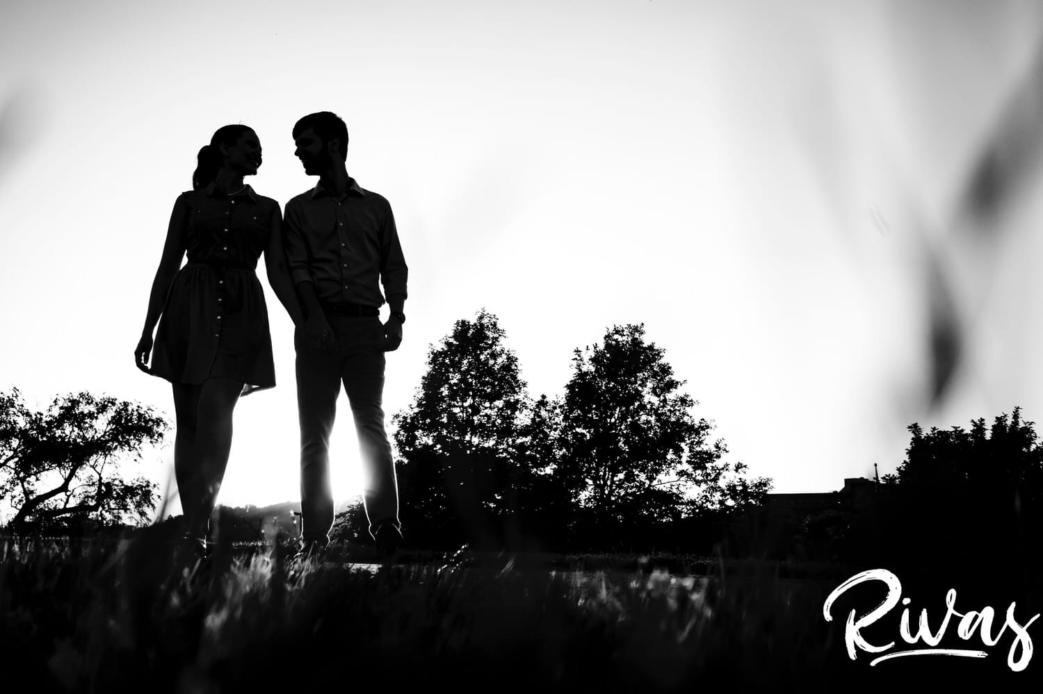 A dramatic, black and white portrait of a couple holding hands and leaning in towards each other, silhouetted against the setting sun during their KC engagement session. 