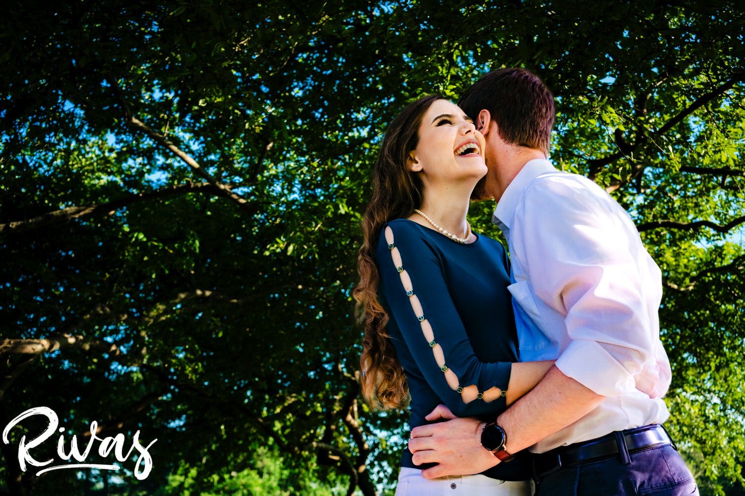 A colorful, candid picture of a man whispering in his finace's ear as they stand underneath a bright green canopy of trees during their engagement session at Kansas City's Nelson Atkins Museum of Art. 