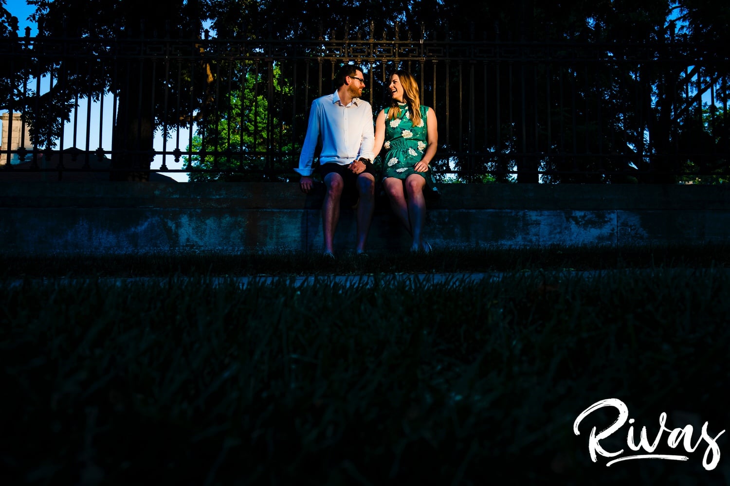 A vibrant picture taken from a distance of an engaged couple sitting on the edge of a wrought iron fence at sunrise during their engagement with their wedding photographer in Kansas City. 
