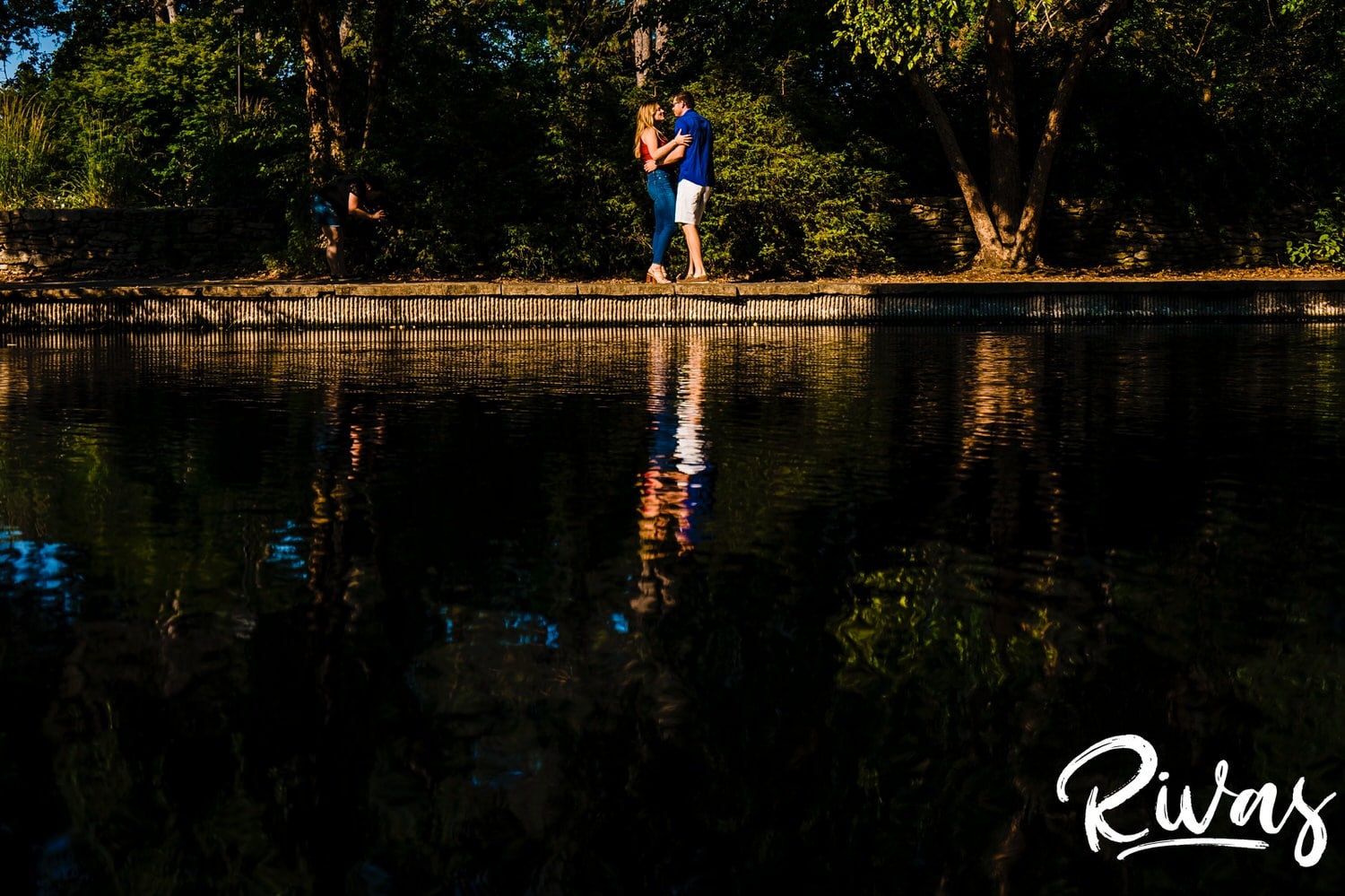 A very wide, colorful picture taken across a lake of a couple dancing together during their summer engagement session, their reflections visible in the lake. 