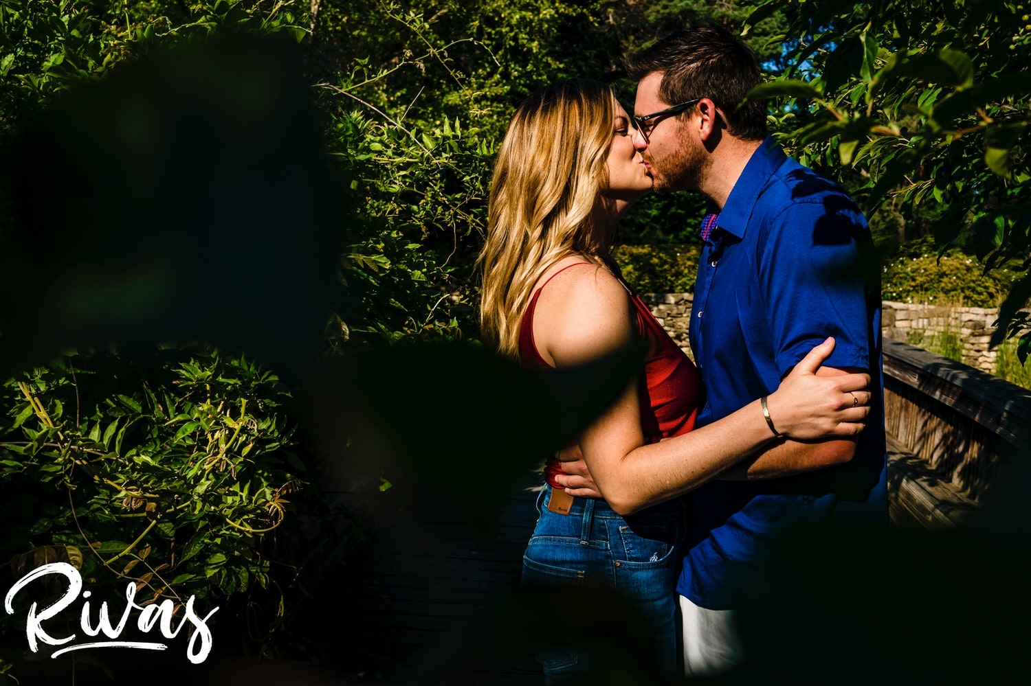 A colorful, intimate portrait of an engaged couple sharing a kiss underneath a canopy of bright green leaves during their engagement session with their wedding photographer. 