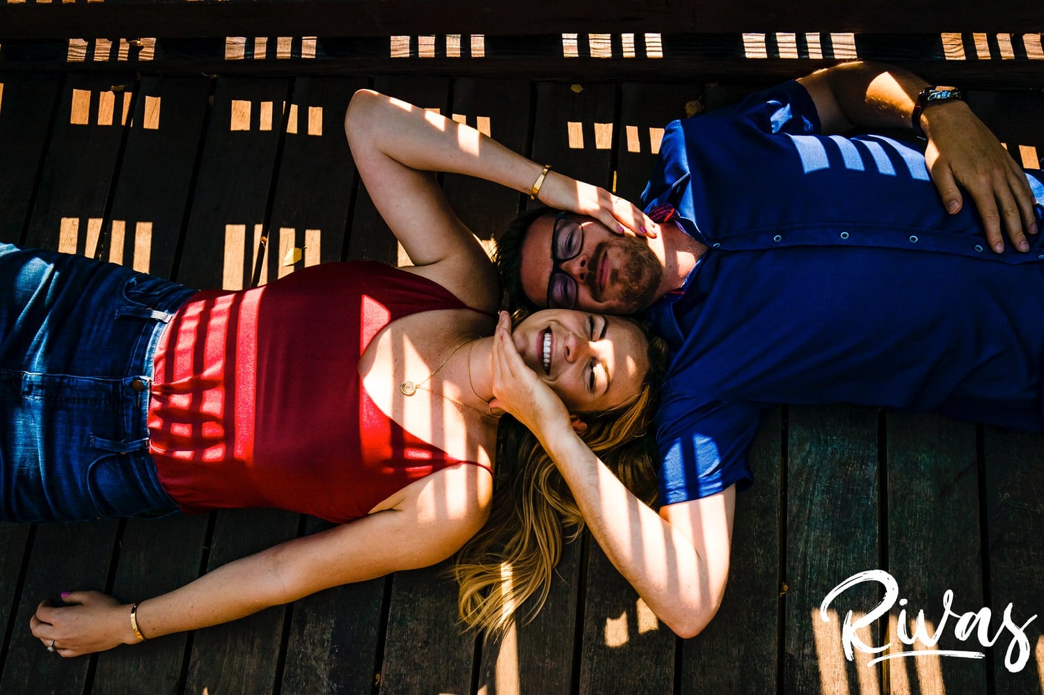 A silly, candid picture of an engaged couple lying on a wooden bridge, with sunlight creating a pattern on their faces as they hold each others' face, laughing hysterically during their summer engagement session at Loose Park in Kansas City. 