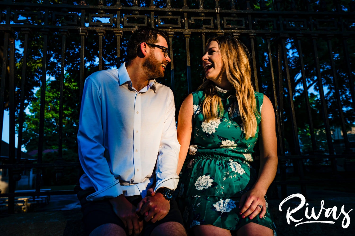 A vibrant picture taken from a distance of an engaged couple sitting on the edge of a wrought iron fence at sunrise during their engagement with their wedding photographer in Kansas City. 