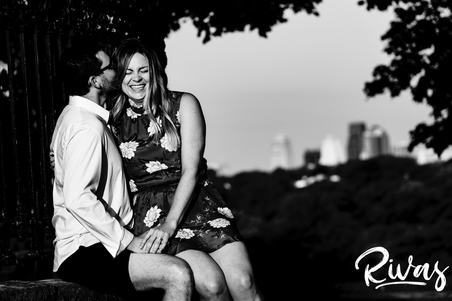 A candid black and white picture of a man whispering into his fiance's ear with the Kansas City skyline visible in the background. 