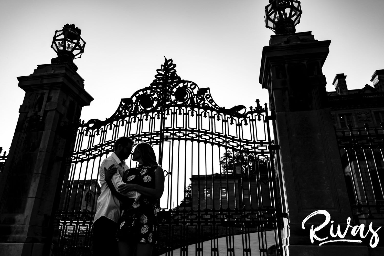 A dramatic black and white portrait of an engaged couple wrapped up in each others' arms as they stand in front of a large, ornate gate during their engagement session with a wedding photographer in Kansas City. 