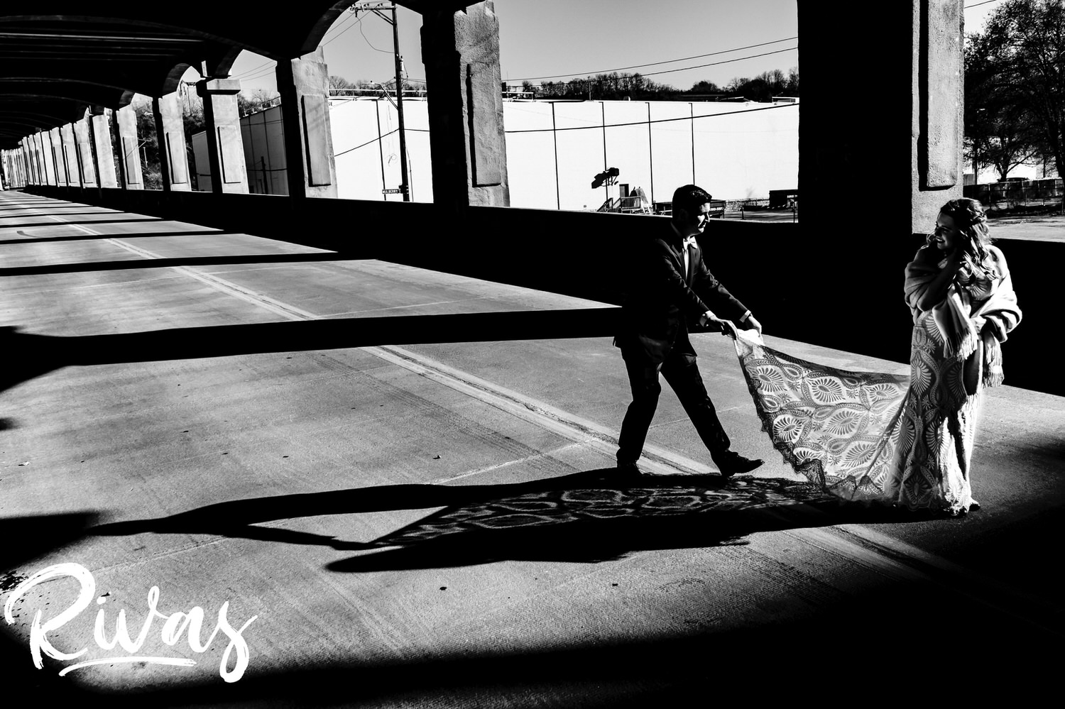 A playful black and white picture of a groom fluffing his bride's train, their shadows visible on the street beside them on the afternoon of their fall wedding reception at The Foundation Event Space in Kansas City. 
