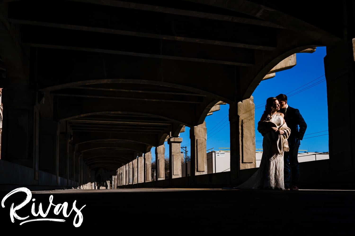 A vibrant, wide picture of a bride and groom leaning in for a kiss as they stand in a splash of sunlight under the 12th Street Viaduct on their fall wedding reception day at The Foundation Event Space in Kansas City. 