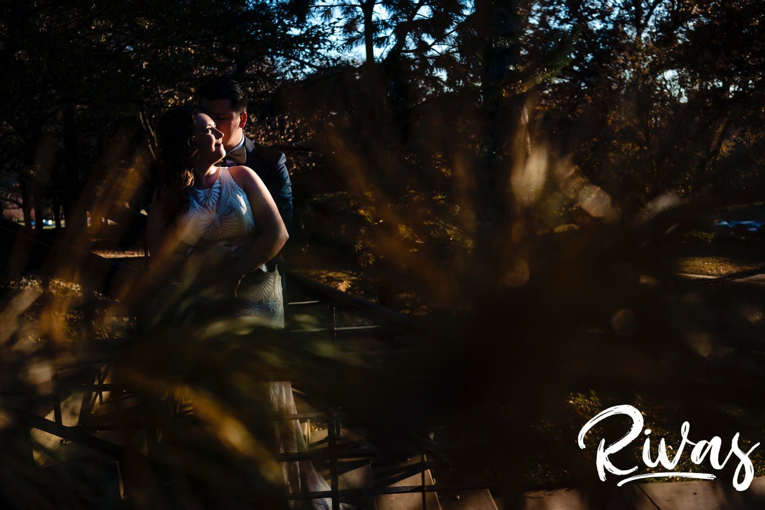 A colorful portrait taken through the trees of a groom leaning in to kiss his bride on the cheek on the afternoon of their fall wedding reception at The Foundation Event Space in Kansas City. 