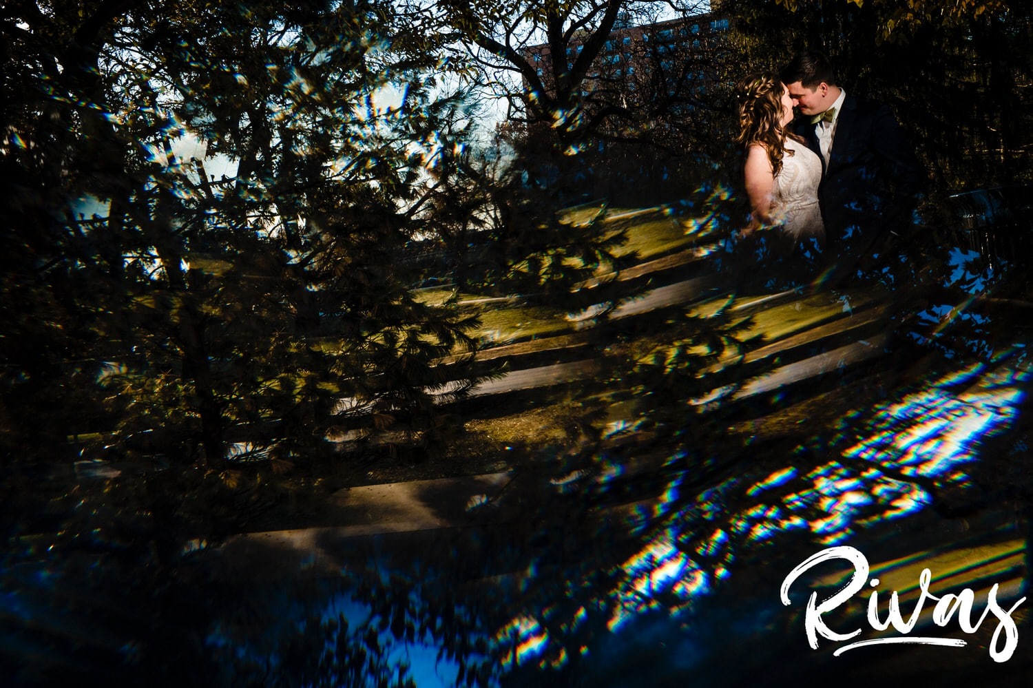 A colorful portrait taken through the trees of a groom leaning in to kiss his bride on the cheek on the afternoon of their fall wedding reception at The Foundation Event Space in Kansas City. 