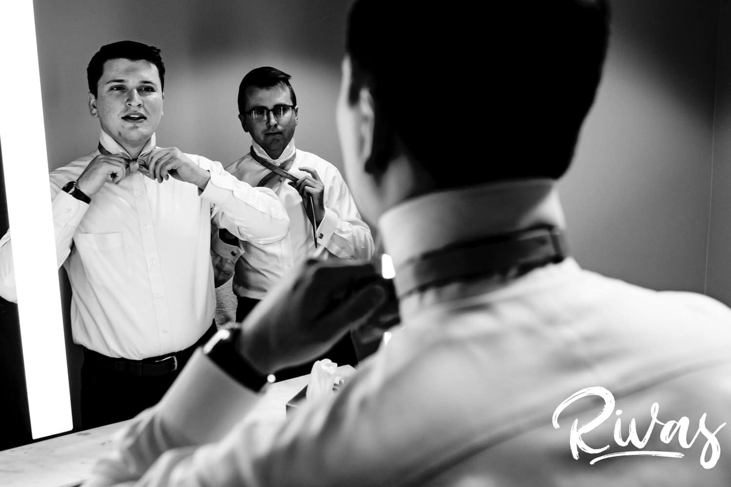 A candid black and white picture of a groom attempting to tie his bow tie in a mirror next to his brother on the morning of his fall wedding reception at The Foundation Event Space in Kansas City. 