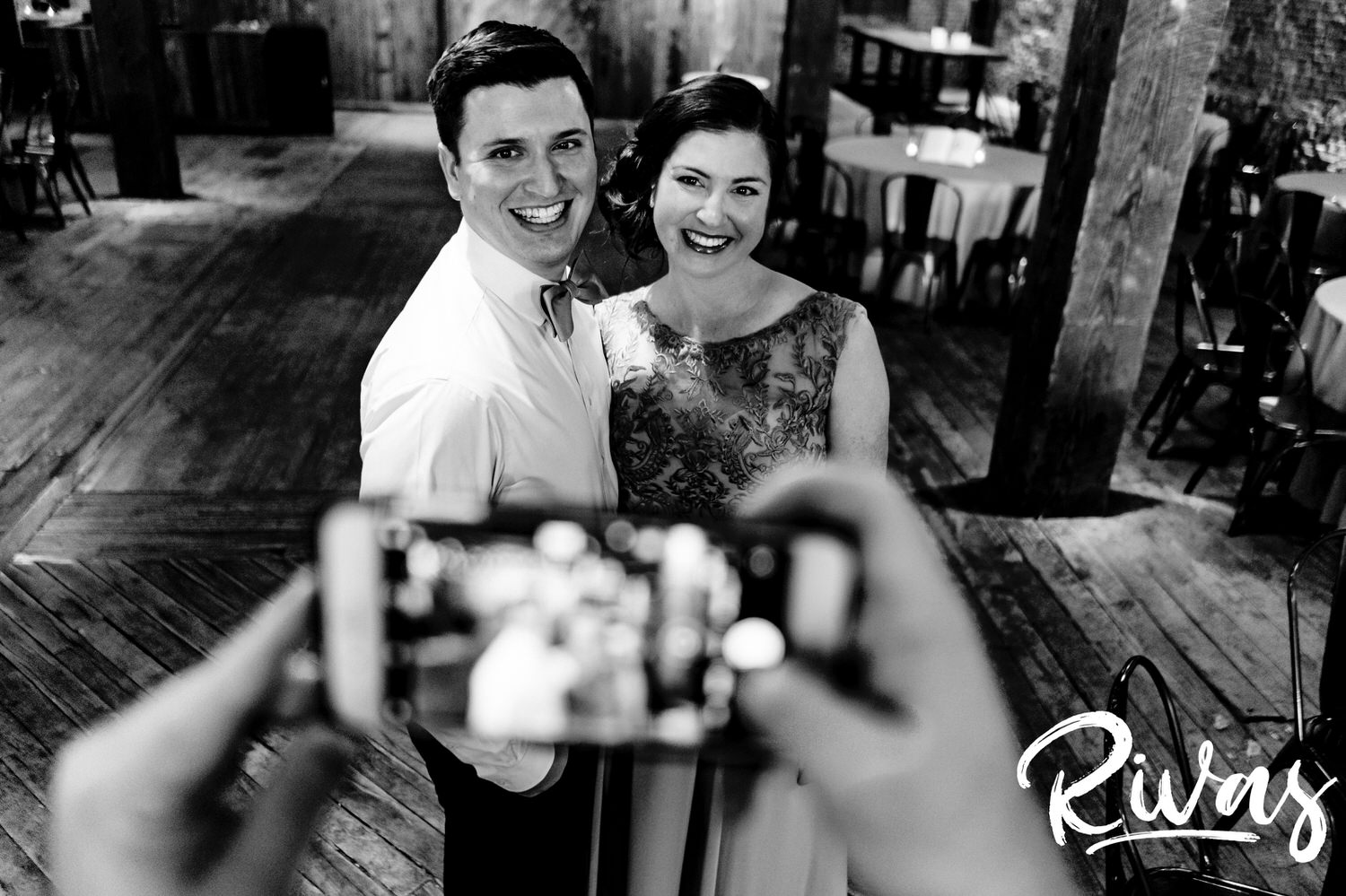 A candid black and white picture taken over the shoulder of a woman taking a photo with her cell phone of a groom and his sister on the afternoon of his fall wedding day. 