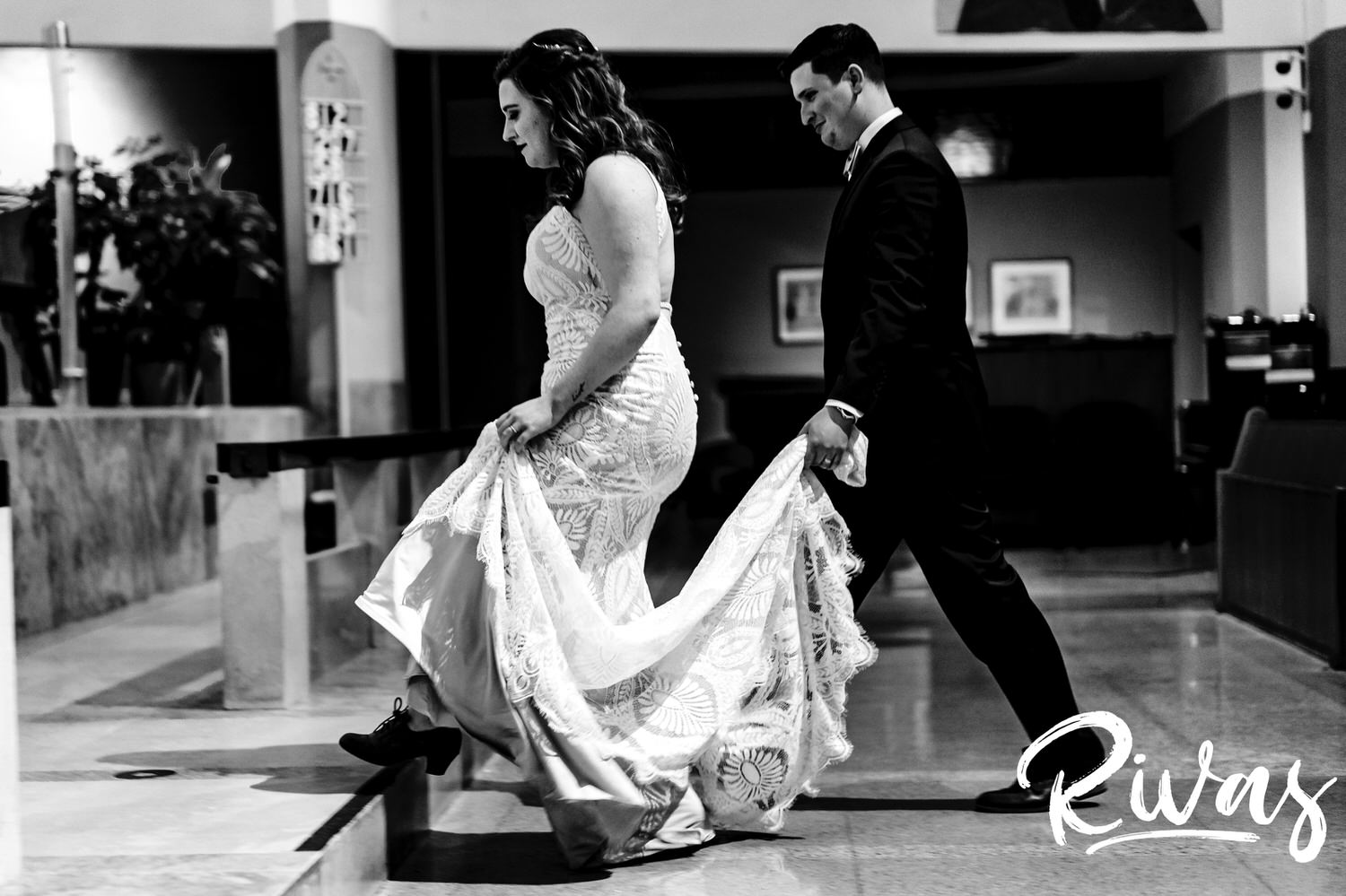 A candid black and white picture of a groom carrying his bride's train up a set of stairs during their wedding ceremony in Kansas City. 