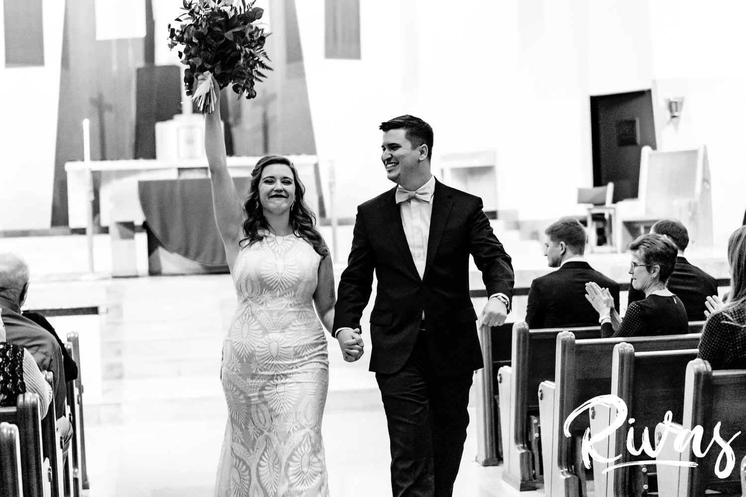 A candid black and white picture of a bride raising her arms in celebration as her groom smiles and laughs as they walk back up the aisle at the end of their wedding ceremony in Kansas City. 