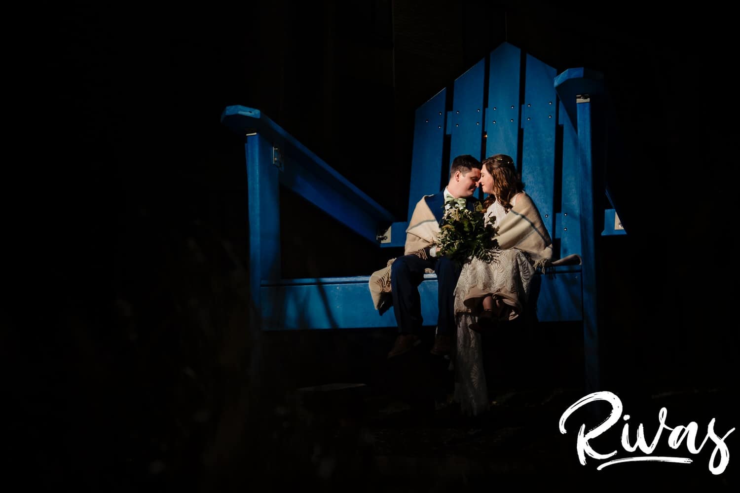 A colorful photo taken at night of a bride and groom snuggling in and sharing a kiss as they sit on a giant, blue, Adirondack chair on the campus of Rockhurst University on the evening of their fall wedding in Kansas City. 