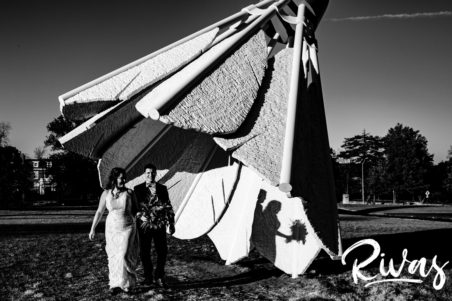A candid black and white picture of a bride and groom holding hands and smiling as they walk away from a giant shuttlecock on the back lawn of the Nelson on their fall wedding day in Kansas City. 