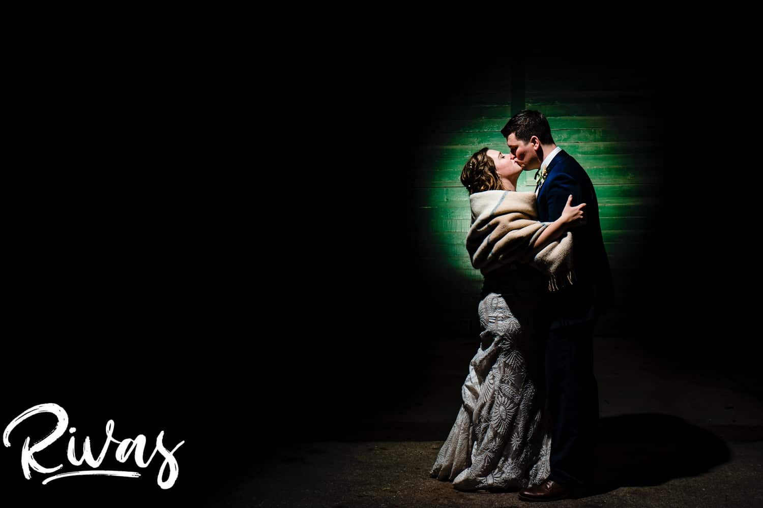 A colorful portrait of a bride and groom, spotlit against a green wall sharing a kiss at the end of their fall wedding reception at The Foundation Event Space in Kansas City. 