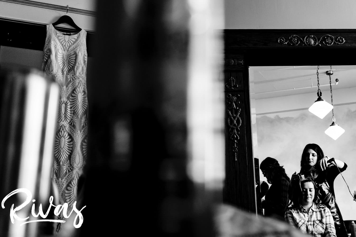 A candid black and white picture of a bride's reflection in a mirror, eyes closed as her hair gets curled with her wedding dress visible on the left side of the frame on her fall wedding day at The Foundation Event Space in Kansas City. 