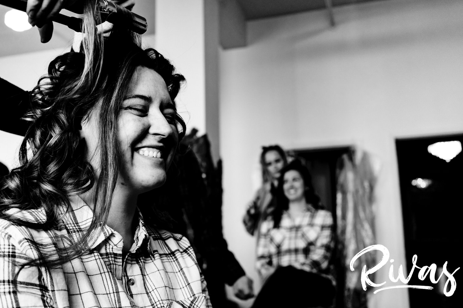 A candid black and white picture of a bride laughing with a bridesmaid visible in the background as she gets her hair curled on the day of her fall wedding reception at The Foundation Event Space in Kansas City. 