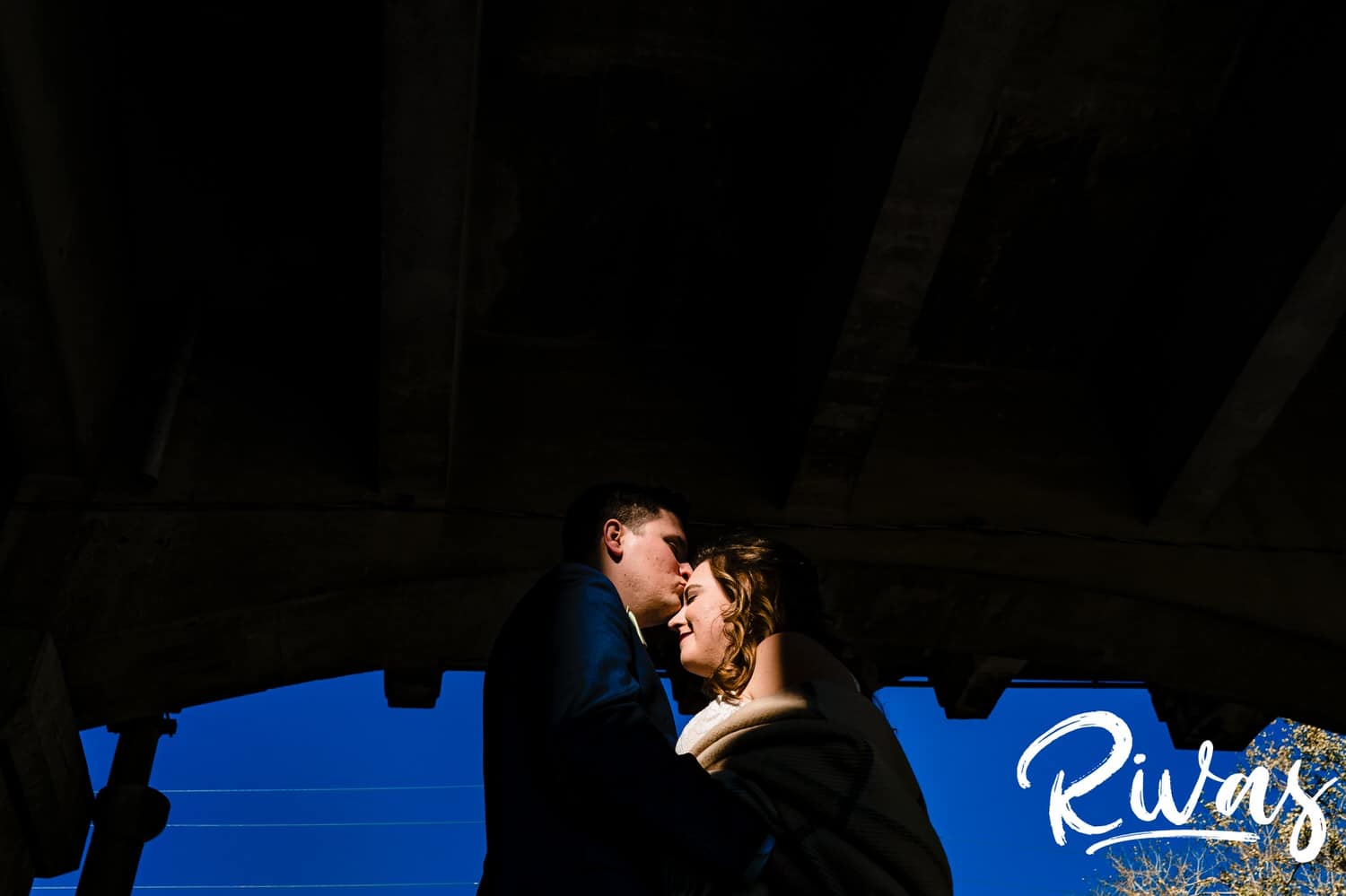 A vibrant, wide picture of a bride and groom leaning in for a kiss as they stand in a splash of sunlight under the 12th Street Viaduct on their fall wedding reception day at The Foundation Event Space in Kansas City. 