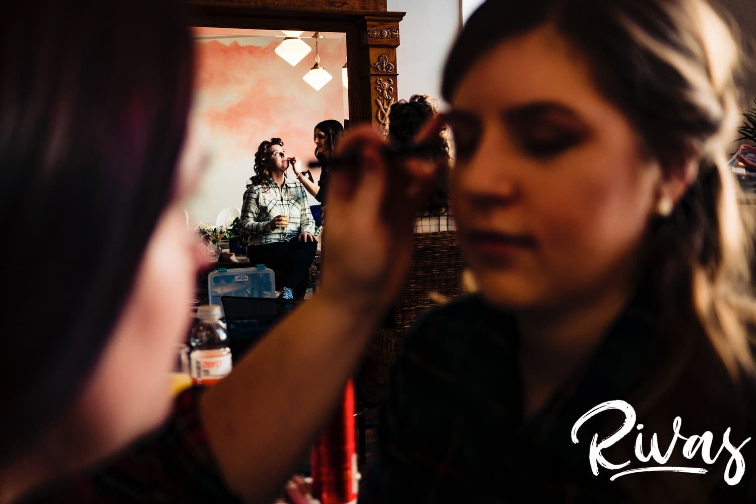 A warm, colorful photo of a bride getting her makeup done in the background as a bridesmaid gets her makeup done in the foreground on the morning of a fall wedding reception at The Foundation Event Space in Kansas City. 