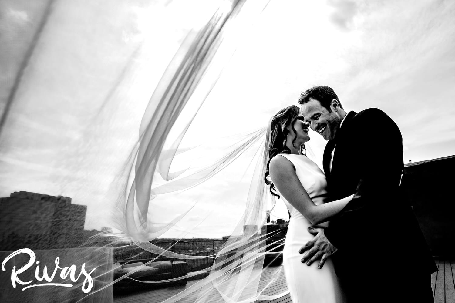A candid black and white portrait of a bride and groom sharing an embrace and laughing together as the bride's veil blows in the wind just after their first look on their KC Skyline wedding day. 