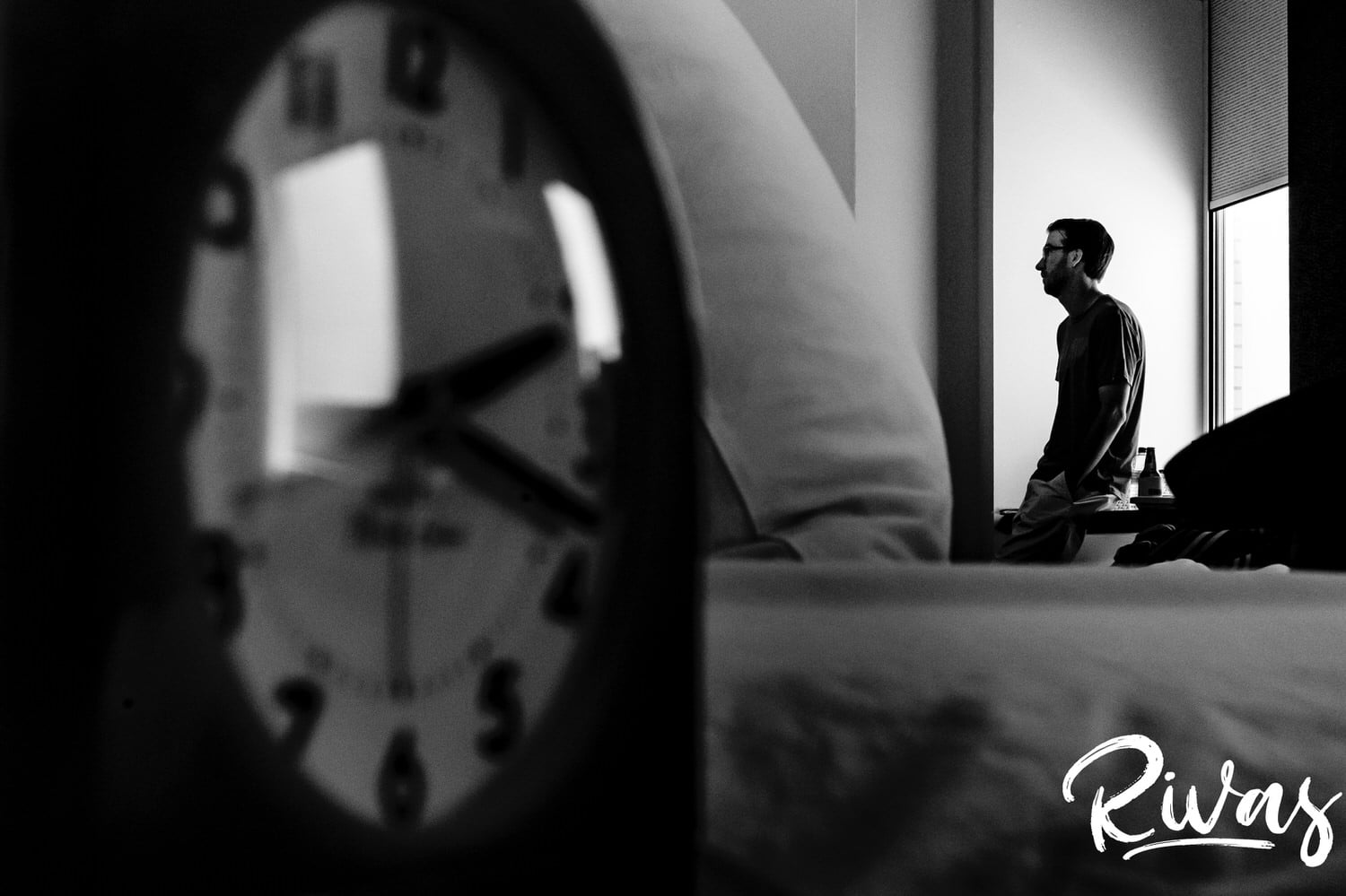 A black and white picture of the face of a clock showing 2:18 in the foreground, with a groom visible in the background starting to get dressed on the afternoon of his Leawood wedding. 