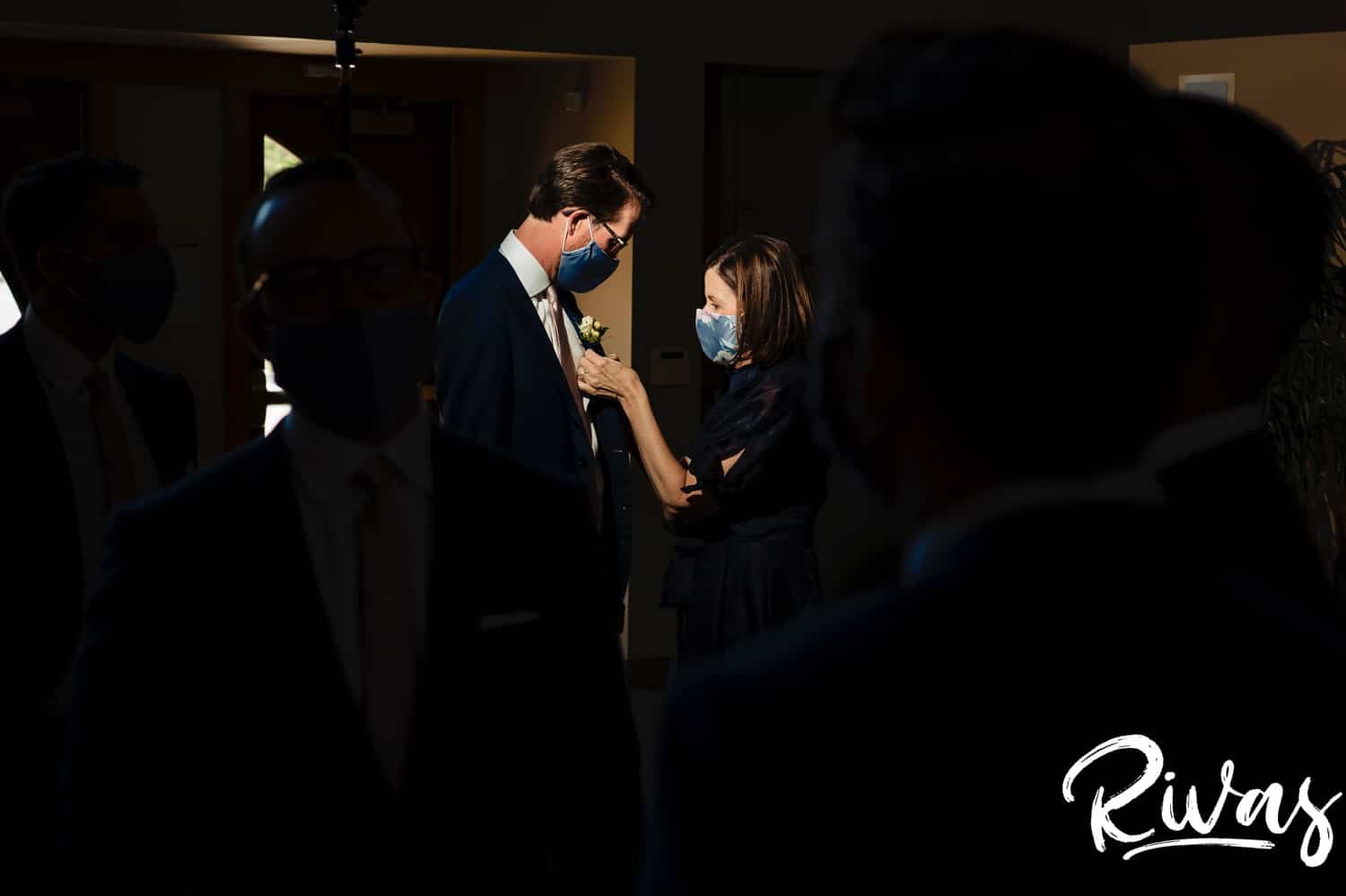 A candid picture taken from a distance of a groom's mom putting his bouttioniere on, both in masks on the afternoon of his wedding reception at The Museum at Prairiefire. 