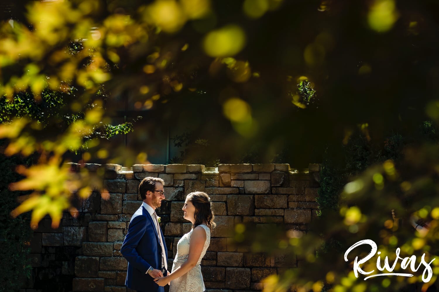 A colorful portrait taken through a canopy of bright green trees of a bride and groom holding hands, looking at each other just after their first look on their wedding day in Overland Park, Kansas. 