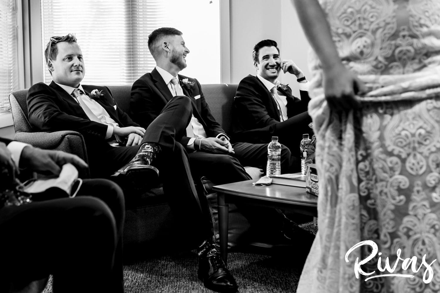 A candid black and white picture of a bride holding her gown and walking away from a groom of groomsmen sitting on a couch laughing just before a wedding ceremony at Wesley Chapel in Overland Park. 