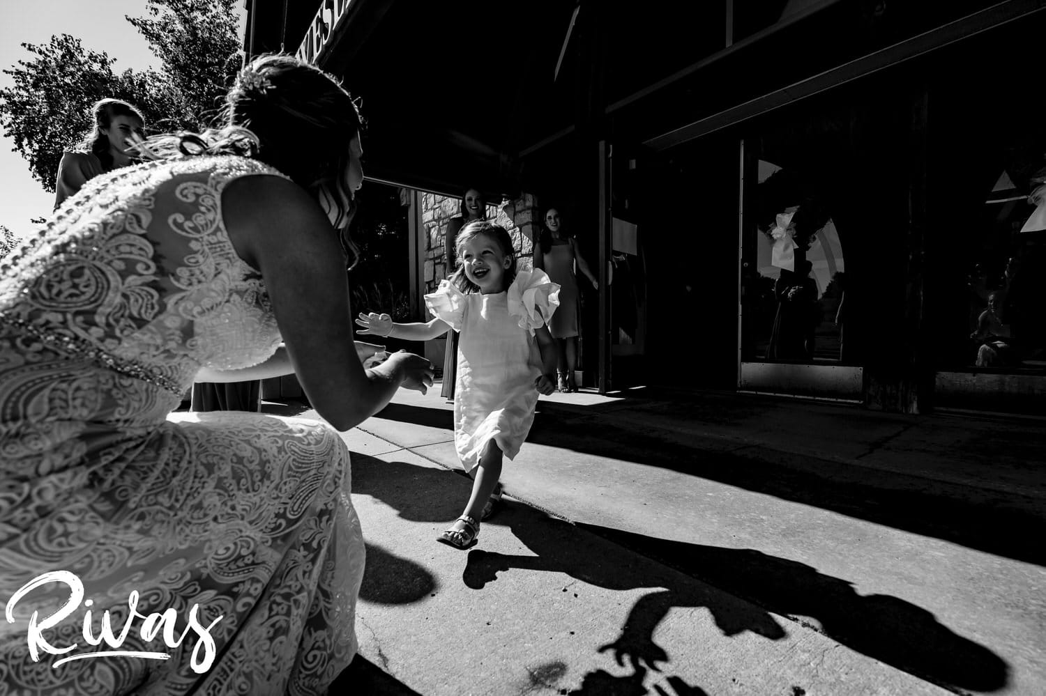 A candid black and white picture of a flower girl with ruffly sleeves running towards a bride, arms outstretched on the afternoon of a wedding in Leawood, Kansas. 