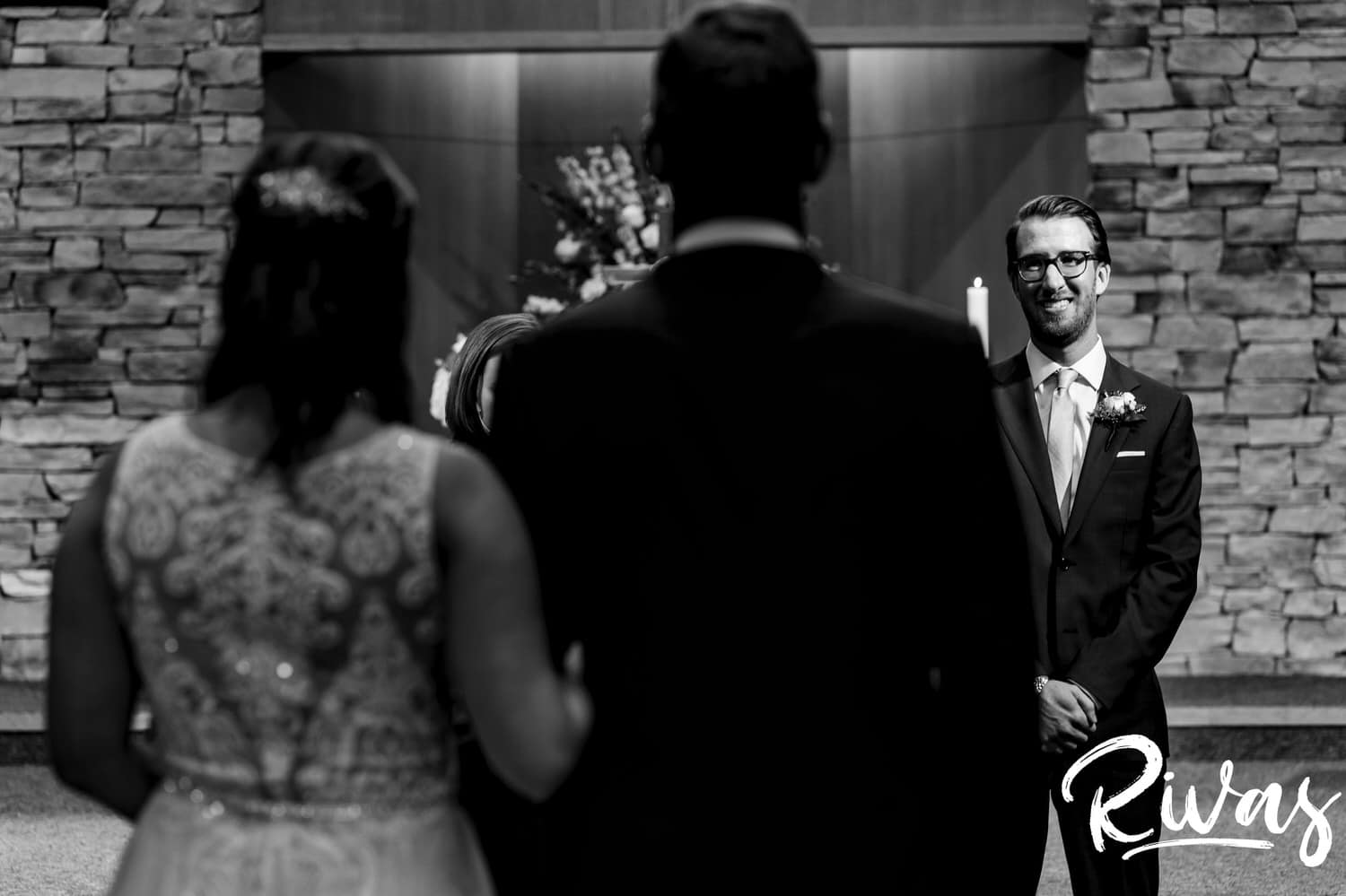 A candid black and white picture taken over the shoulder of a bride and her father of her groom smiling at her as she walks down the aisle during their wedding ceremony in Kansas City. 