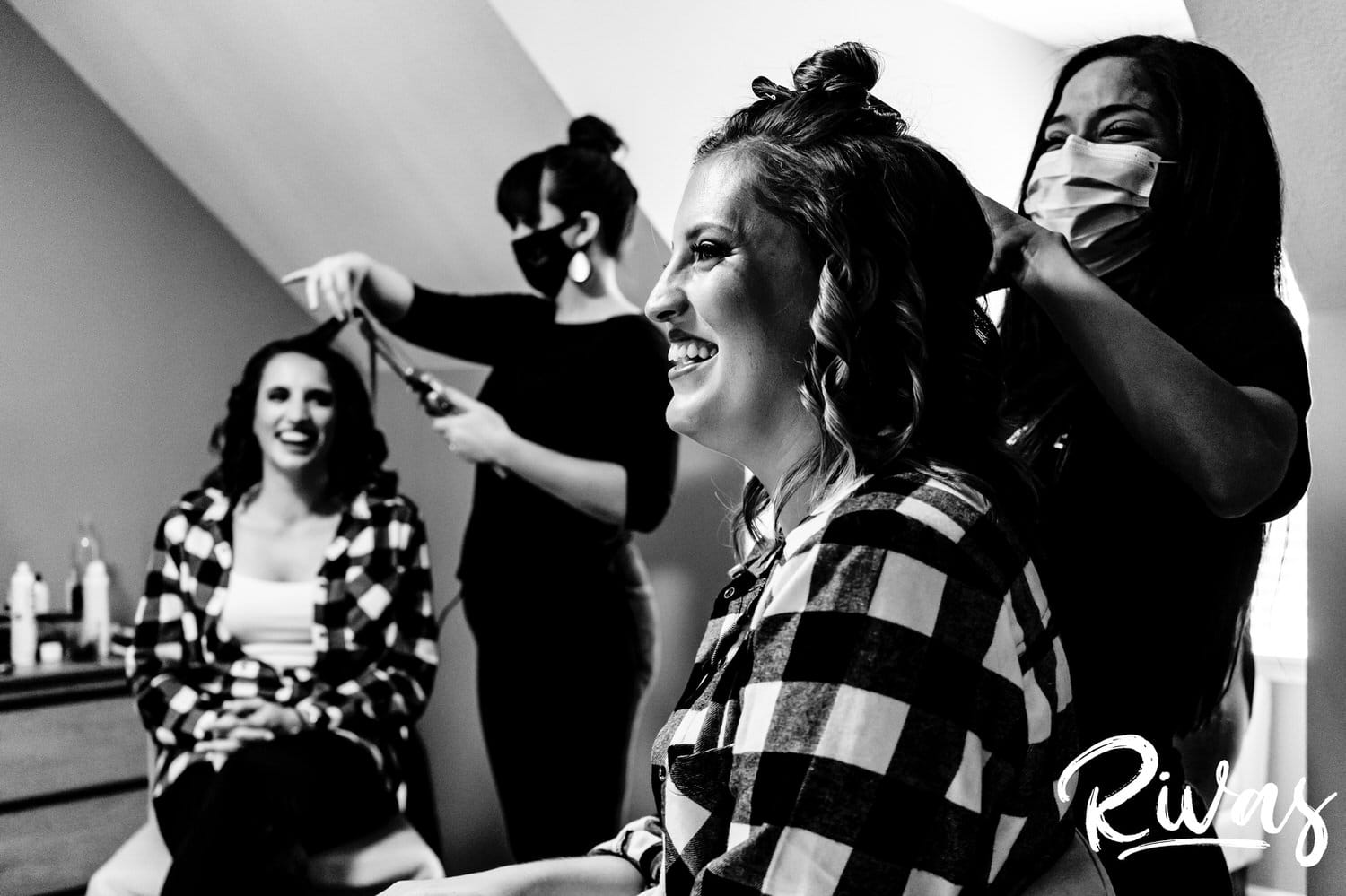 A bold black and white candid picture of a bride in a black and white plaid shirt getting her hair curled as she laughs with a bridesmaid on the morning of her wedding in Kansas City. 