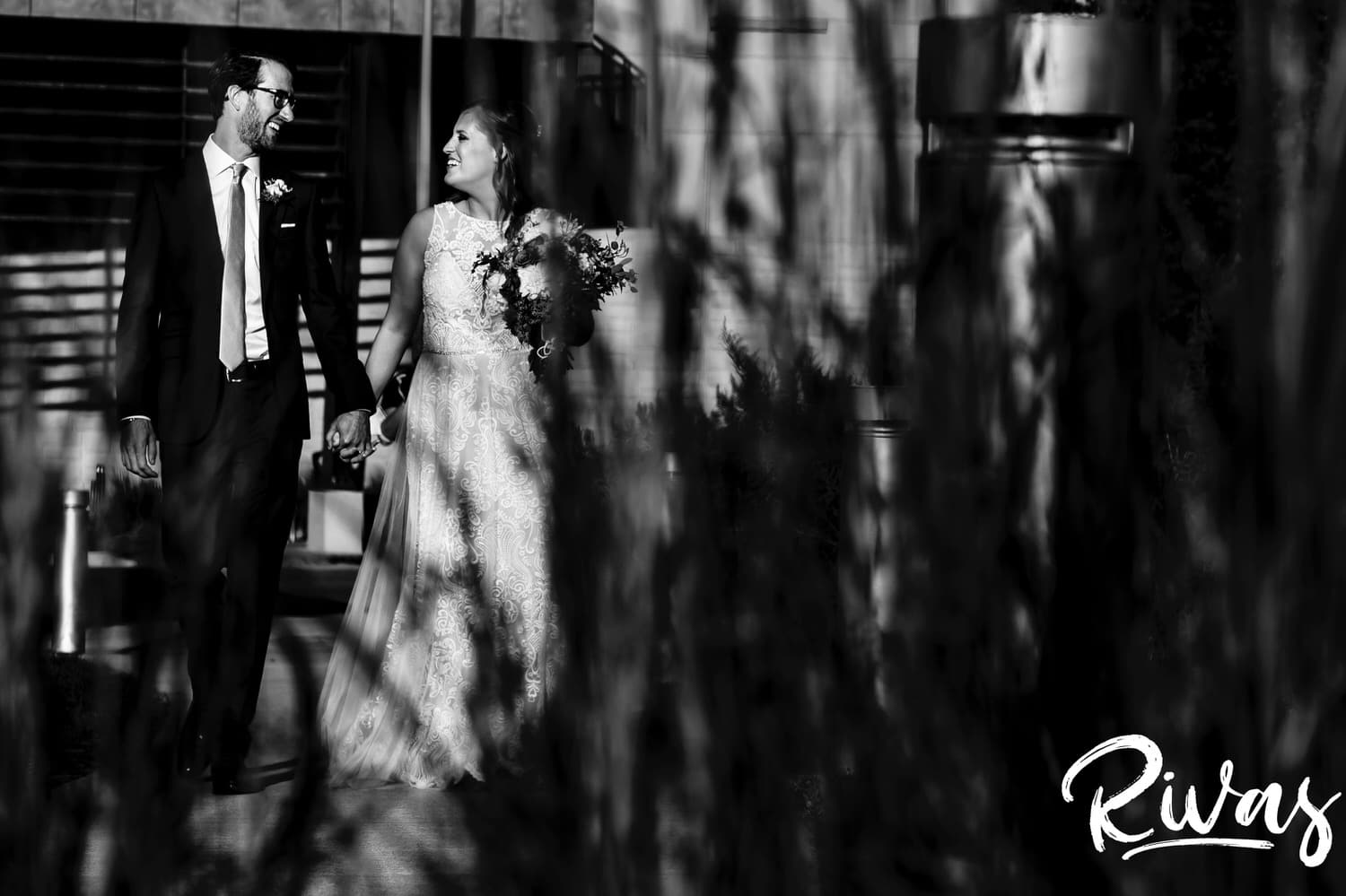 A black and white picture taken through a clump of grass of a bride and groom holding hands, walking into the sunset on the day of their Museum at Pairiefire Wedding reception. 