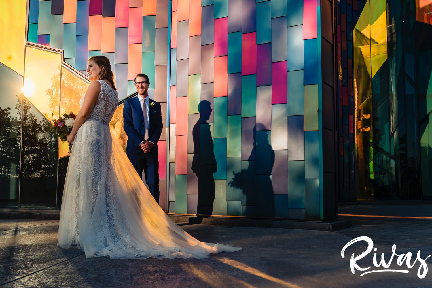 A colorful picture of a bride and groom holding hands, laughing together as they stand in front of the colorful tiled wall at The Museum at Prairiefire on the evening of their wedding. 