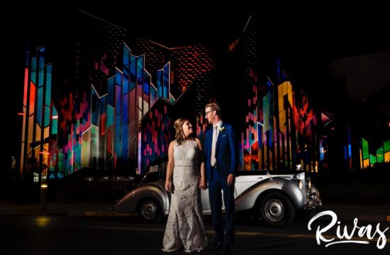 A vibrant picture of a bride and groom holding hands and looking at each other as they stand in front of an antique Bentley parked in front of the Museum at Prairiefire on their wedding night in Kansas City.